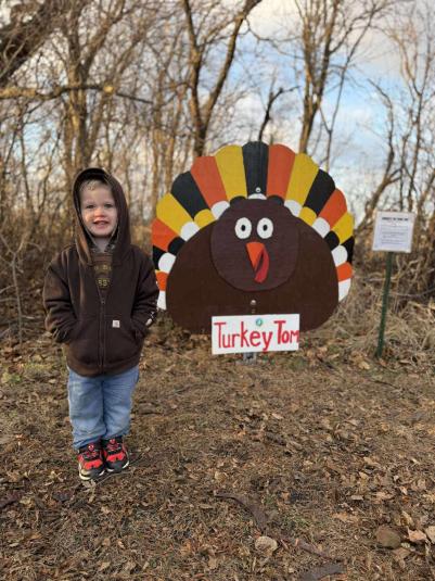 A young boy, around 3 years of age, stands in front of a brown, orange, and yellow wooden turkey