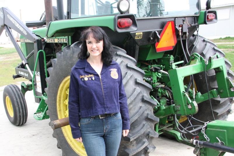 Darcy Dougherty Maulsby stands in front of a green tractor wearing her FFA jacket. 