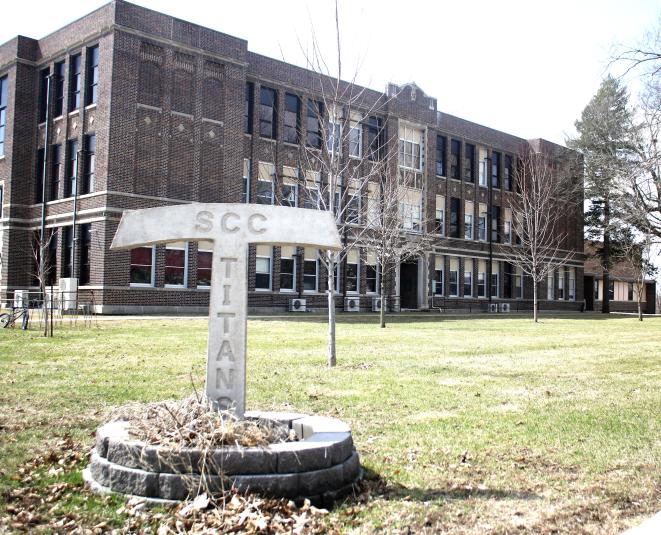 The exterior of the SCC High School, a three-story brick building. In the foreground is a sign in the shape of the letter "T" with the word "Titan" engraved on it. 