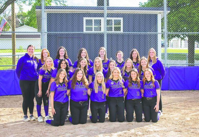 Members of the South Central Calhoun softball team pose with their coaches. They are outside on a sunny day, standing on the softball field. The players are wearing purple jerseys and black pants.